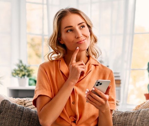 woman sitting cross-legged on a couch in a bright sunny room holding her phone in one hand and putting her index finger up to her chin as if she's thinking. 